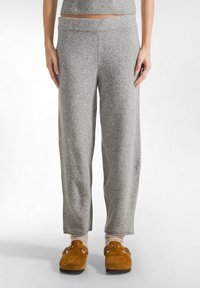 Grijze, zachte, wijde sweatpants met een brede tailleband; gedragen met bruin suède pantoffels en crèmekleurige sokken. Minimalistisch ontwerp.