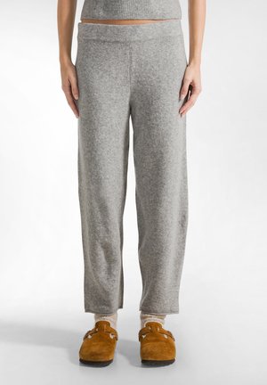 Grijze, zachte, wijde sweatpants met een brede tailleband; gedragen met bruin suède pantoffels en crèmekleurige sokken. Minimalistisch ontwerp.