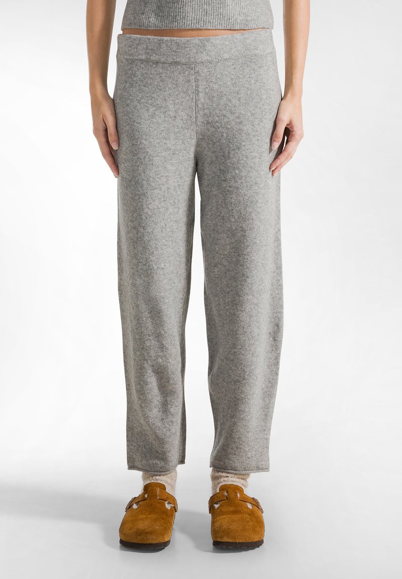 Grijze, zachte, wijde sweatpants met een brede tailleband; gedragen met bruin suède pantoffels en crèmekleurige sokken. Minimalistisch ontwerp.