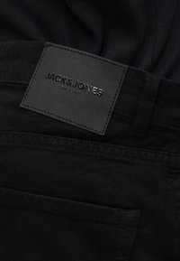 Μαύρο τζιν από ντένιμ με ραμμένο ορθογώνιο δερμάτινο ταμπελάκι που γράφει "JACK&JONES SINCE 1990" κοντά στη μέση και τη θηλιά της ζώνης.