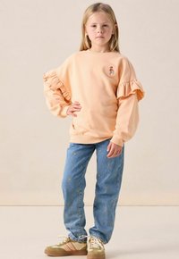 Meisje staat met hand op de heup, draagt een perzikkleurige sweatshirt met ruches, blauwe jeans met gouden stippen en beige sneakers met roze strepen.