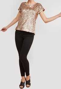 Top z cekinami w kolorze różowego złota z krótkimi rękawami i luźnym krojem, zestawiony z czarnymi skinny jeansami i czarnymi sandałami na obcasie.