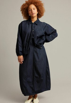 Persoon met krullend haar die een donkerblauw spijkerjack en een lange rok draagt, staand met één hand op de heup en witte schoenen op een effen achtergrond.
