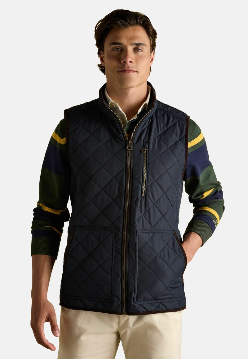 Námorný prešívaný vest s vysokým golierom, predným zipsom, dvoma prednými vreckami a hnedými detailmi. Nosí sa cez svetlý sveter so zelenými a modrými pruhmi.