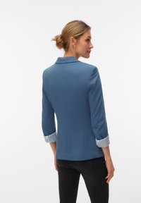 Blauwe blazer met een getailleerde pasvorm, voorzien van een reverskraag en opgerolde gestreepte manchetten. Gladde stof met een subtiele glans. Zwarte jeans zichtbaar.