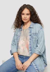 Denim-Hemd mit Knöpfen, hellblau, über einem grauen T-Shirt mit grafischem Sonnenstrahl-Design getragen. Modell in lässigen blauen Jeans, in sitzender Pose.