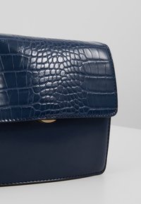 Mørkeblå faux krokodille læder håndtaske med glat overflade, struktureret form, fold-over klap og hardware i guldtoner.