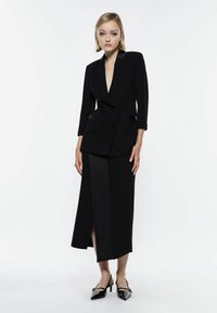 Blazer nero su misura con scollo a V profondo, design doppiopetto e rever in satin, abbinato a una gonna maxi coordinata con uno spacco frontale.