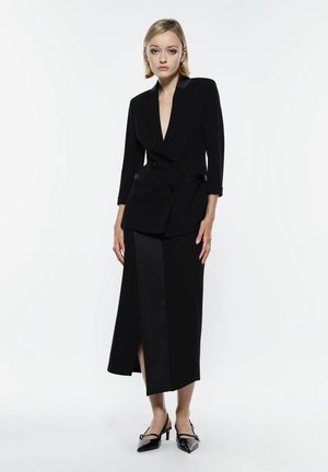 Blazer nero su misura con scollo a V profondo, design doppiopetto e rever in satin, abbinato a una gonna maxi coordinata con uno spacco frontale.