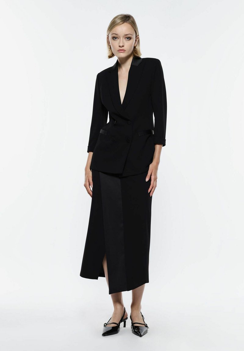 Blazer nero su misura con scollo a V profondo, design doppiopetto e rever in satin, abbinato a una gonna maxi coordinata con uno spacco frontale.