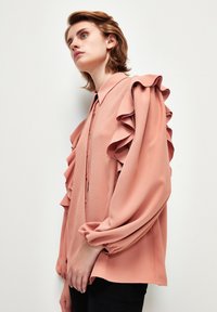 Blusa rosa con mangas acampanadas, cierre de botones en el frente y cuello. La tela tiene una textura sutil, las mangas son abullonadas y están fruncidas en los puños.