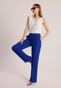 Niebieskie spodnie wide-leg z gładkiego materiału, zestawione z białym topem bez rękawów z dekoltem w kształcie litery V i przezroczystymi akcentami.