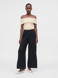 Femme portant un haut crème à épaules dénudées, pantalon noir à jambes larges et escarpins blancs à bouts pointus, debout devant un fond clair.