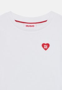 Άσπρο βαμβακερό T-shirt με στρογγυλή λαιμόκοψη, που διαθέτει ένα μικρό κόκκινο μπαλώμα σε σχήμα καρδιάς με κεντημένο το "HUGO" σε λευκό κείμενο. Εσωτερική λαιμόκοψη με κόκκινη λεπτομέρεια.
