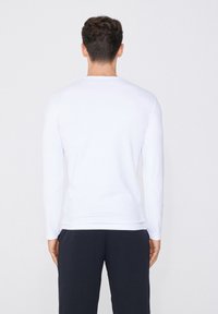 Camisa blanca de manga larga hecha de una tela suave, con cuello redondo y corte relajado, combinada con pantalones oscuros, vista desde atrás.