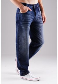 Herren Jeans aus blauem Denim mit geradem Schnitt, leichten Verwaschungen und gebürsteter Textur. Getragen mit weißen Sneakers mit roten Akzenten.