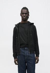 Sudadera con capucha negra con cremallera y bolsillo frontal, combinada con una camiseta negra y vaqueros azul oscuro. La sudadera tiene cordones y detalle de logo.