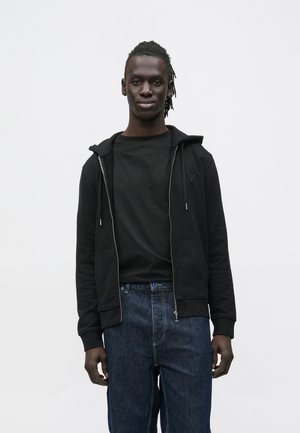 Felpa nera con cerniera e tasca frontale, abbinata a una t-shirt nera e jeans blu scuro. La felpa presenta coulisse e dettaglio con logo.