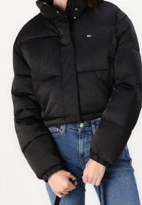 Czarna krótka kurtka puchowa z pikowanym wzorem, wysokim kołnierzem i zapięciem na napy. Materiał wydaje się błyszczący i gładki. Noszona na niebieskich dżinsach.