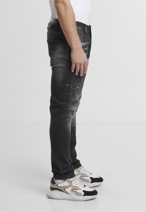 Homme debout de profil, portant un jean noir délavé avec des éclaboussures de peinture et des baskets blanches, beiges et noires.