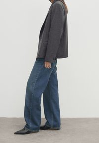 Blazer grigio, strutturato, con taglio ampio sopra jeans di denim blu a vestibilità morbida. Le scarpe sono nere e appuntite con una finitura liscia. Design minimalista.