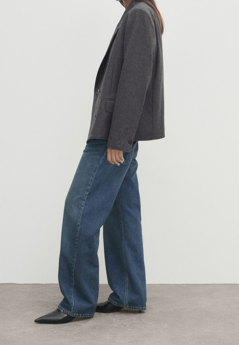 Blazer grigio, strutturato, con taglio ampio sopra jeans di denim blu a vestibilità morbida. Le scarpe sono nere e appuntite con una finitura liscia. Design minimalista.