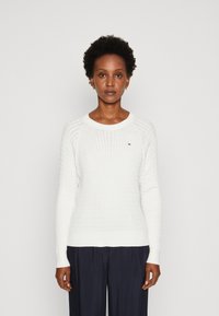 Maglione bianco a maglia con texture a coste, scollo rotondo e maniche lunghe. Presenta un piccolo logo sul petto e un orlo aderente.