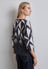 Frau, die ein langärmliges Oberteil mit schwarz-weißer geometrischer Musterung und dunkelblauen Jeans trägt, seitlich an einer schlichten hellen Wand steht.