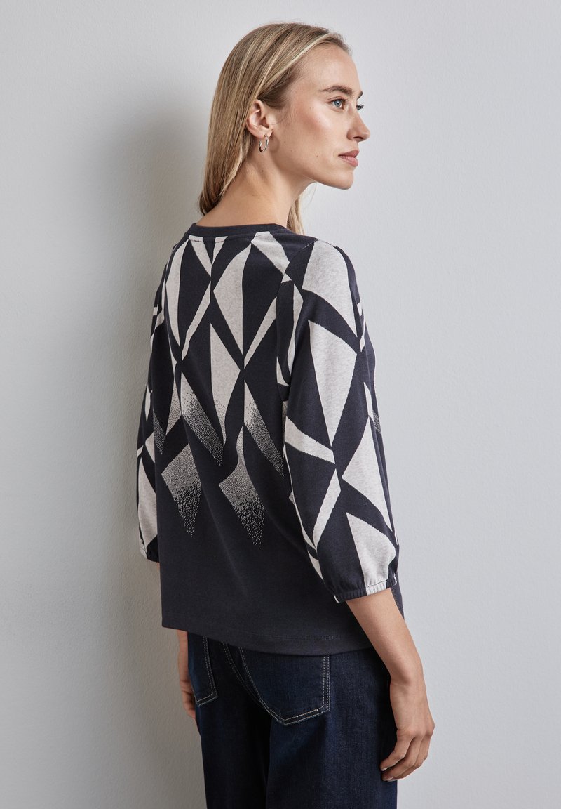 Frau, die ein langärmliges Oberteil mit schwarz-weißer geometrischer Musterung und dunkelblauen Jeans trägt, seitlich an einer schlichten hellen Wand steht.