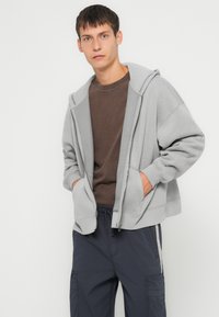 Felpa grigia con zip e un ampio cappuccio, tasche frontali e vestibilità rilassata, indossata sopra una maglia marrone e abbinata a pantaloni cargo blu navy.