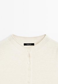 Cardigan couleur crème avec col rond et une rangée de six boutons sur le devant, arborant une étiquette Massimo Dutti à l'intérieur du col.