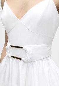 Λευκό φόρεμα χωρίς μανίκια με βαθύ V-ντεκολτέ και φαρδιά ζώνη με ρυτιδώσεις που διαθέτει ορθογώνια αγκράφα σε χρυσό τόνο στη μέση.