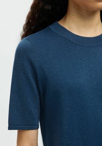 Lyhythihainen sininen neulepaita, jossa on tekstuuripinta. Paidan rinnassa on ribbikuvioinen kaula-aukko ja käänteet hihoissa, ja se tarjoaa hienovaraisen kiillon.