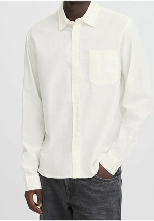 Homme portant une chemise blanche à manches longues avec poche poitrine et un jean gris foncé, une main dans la poche, fond uni.