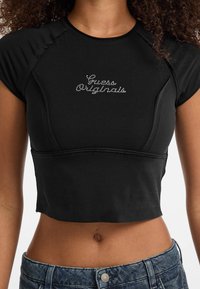 Zwarte cropped top van zachte stof, met korte mouwen, een strakke pasvorm en "Guess Originals" geschreven in rhinestones op de voorkant.