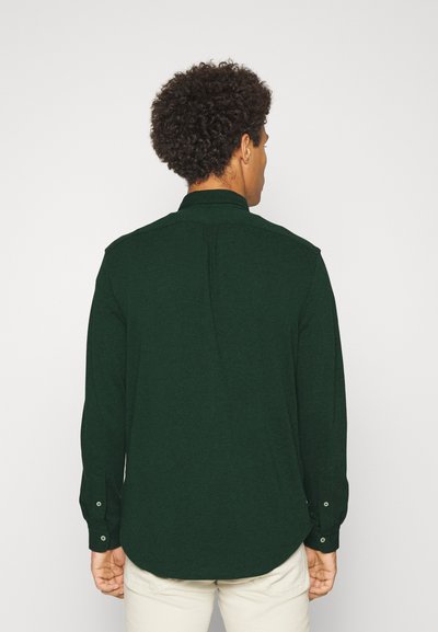 Donkergroen shirt met lange mouwen en een afgeronde zoom, met knoopmanchetten, een rugpas en een gestructureerde stof. Draagt over lichtgekleurde broek.