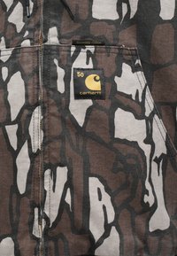 Canvasjacke mit einem braun-grauen abstrakten Muster, Carhartt-Logo-Patch und goldenen Jubiläumsdetails auf der Brusttasche.