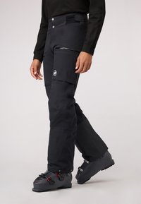 Zwarte skibroek met een slim fit, voorzien van een zijzak, waterdicht materiaal en versterkte onderkanten. Gedragen met zwarte skischchoenen.