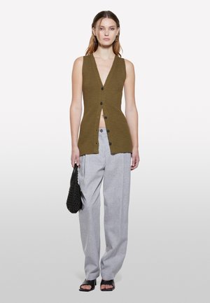 Olivgrüner ärmelloser Rippenstrick-Cardigan mit tiefem V-Ausschnitt, vorderer Knopfleiste und Split-Design, kombiniert mit hellgrauen Hosen.