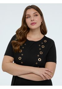 Mujer con cabello largo y castaño, vestida con una blusa negra con adornos circulares dorados, brazos cruzados, mirando ligeramente de lado contra un fondo blanco.