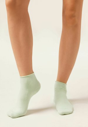 Jambes portant des chaussettes courtes vert pâle, debout sur un sol clair devant un fond neutre.