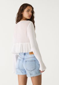 Weißes, strukturiertes Crop-Top mit langen Ärmeln und Rüschensaum, kombiniert mit hellblauen Denim-Shorts mit umgeschlagenen Säumen und Taschen.