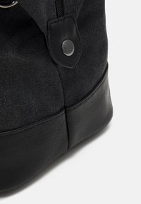 Sac fourre-tout en toile noire, doté d'une base en cuir durable, de textures contrastées, d'une fermeture à bouton-pression et d'un design minimaliste.