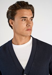 Mörkblå ribbad cardigan med djup V-ringning, enkel knappstängning, bärs över en vit crew-neck t-shirt. Slät textur och figurnära design.