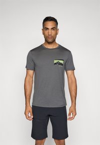 Grå t-skjorte med korte ermer som har et grønt og svart fjellgrafikk-lomme; kombinert med mørke shorts. Glatt stoff, rund hals.