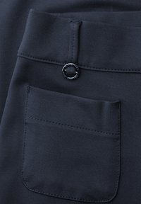 Zbliżenie na kieszeń ciemnych spodni z tkaniny z przeszytą pętlą i małym okrągłym czarnym guzikiem nad kieszenią.