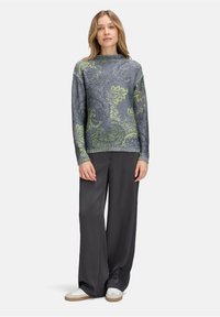 Grauer Pullover mit grünem Paisleymuster, runder Halsauschnitt, lange Ärmel. Kombiniert mit lockeren schwarzen Hosen und weißen Turnschuhen. Glatte Textur.