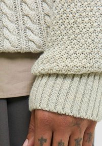 Pull en maille de couleur beige clair avec un motif en câble texturé et des poignets côtelés. Le matériau semble doux et chaud, offrant une sensation de confort.