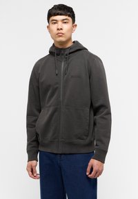 Sudadera con cremallera de color gris oscuro, con capucha ajustable con cordón, dos bolsillos frontales y un logotipo sutil en el pecho. Tejido suave, corte relajado.