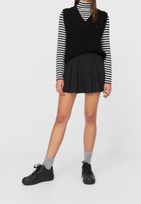 Veste noire en tricot portée sur un chemisier à manches longues rayé noir et blanc, associée à une mini-jupe plissée noire et des baskets noires avec des chaussettes grises.